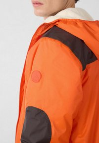 Oranje jas met bruine accenten op schouders en mouwen, voorzien van een kap met zachte voering en een ronde logopatch op de mouw.