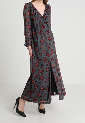 Kvinde i en lang, mørkegrå blomsterkjole med røde blomster, V-hals, lange ærmer, slids foran og sorte højhælede sko.