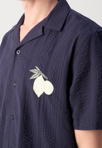 Gros plan d'une personne portant une chemise texturée bleu foncé à manches courtes avec une broderie blanche de citron et de feuille sur la poitrine.