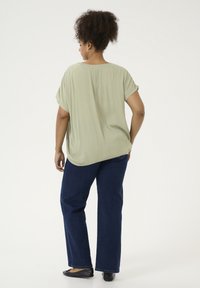 Hellgrünes, kurzärmliges Oberteil aus strukturierter Stoff, kombiniert mit dunkelblauen weit geschnittenen Jeans und schwarzen Ballerinas. Ansicht von hinten.