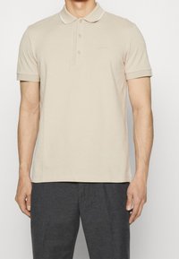 Polo shirt beige à manches courtes avec trois boutons et le logo "BOSS" sur la poitrine gauche, porté avec un pantalon gris foncé.