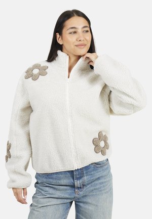 Veste en polaire crème avec un col montant, ornée d'appliqués floraux beiges sur la poitrine et sur le côté, et d'une fermeture éclair sur toute la longueur à l'avant. Texture douce.