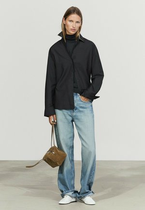 Frau trägt ein schwarzes Hemd über einem schwarzen Top, hellblaue weite Jeans, weiße Schuhe, hält eine braune Wildledertasche am Trageriemen.