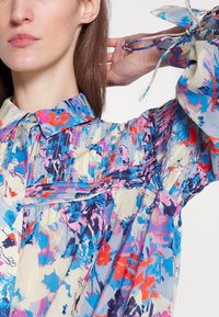 Χρωματιστό φλοράλ μπλουζάκι από ελαφρύ ύφασμα, με γιακά, πτυχωτές λεπτομέρειες και μακριά μανίκια με κορδόνια.