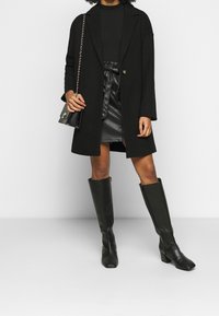 Svart, lång ytterjacka över en svart topp och en kort svart kjol i konstläder. Knähöga svarta stövlar med fyrkantiga tår. Liten crossbody-väska.