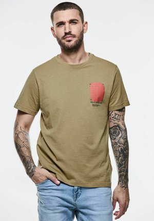 Camiseta de algodón verde oliva con cuello redondo y mangas cortas, que presenta un estampado gráfico en un bolsillo rojo en el lado izquierdo y una textura suave.
