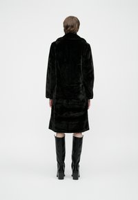 Pessoa de pé, de costas, vestindo um casaco de pelo sintético preto e botas de salto alto até ao joelho pretas, em um fundo branco simples.