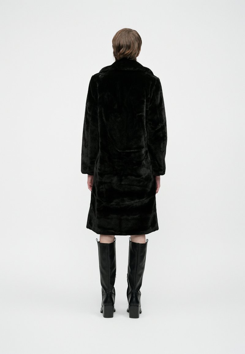 Person steht mit dem Rücken zur Kamera und trägt einen schwarzen Kunstpelzmantel sowie schwarze kniehohe Stiefel mit Absätzen vor einem schlichten weißen Hintergrund.