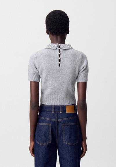 Suéter gris de punto con un cuello adornado con perlas, mangas cortas y una abertura trasera con botones, combinado con jeans oscuros de mezclilla.