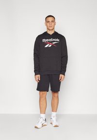 Crna dukserica s Reebok logotipom, prednji džep i kapuljača s vezicom; uparena s tamnim šortsevima i bijelim tenisicama s crnim detaljima.