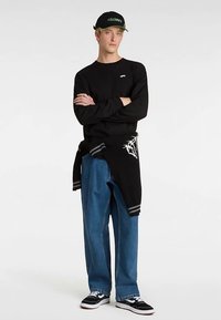 Camiseta negra de manga larga, vaqueros azules de pierna ancha, suéter negro y gris atado a la cintura, gorra de béisbol negra y zapatillas negras.