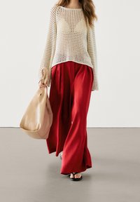 Pull crème léger à maille ouverte, pantalon rouge vif à jambes larges, et un sac fourre-tout beige doux. Textures lisses et contraste de couleurs audacieux.