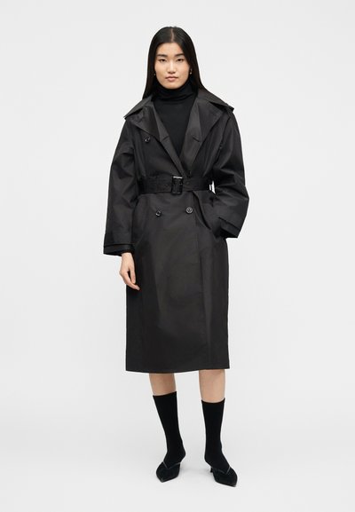 Γυναίκα που φοράει μαύρο trench coat με ζώνη πάνω από μαύρο πουλόβερ με ψηλό γιακά, μαύρες κάλτσες μέχρι το μέσο της γάμπας και μαύρα παπούτσια με μυτερή μύτη, στέκεται μπροστά σε απλο υπόβαθρο.