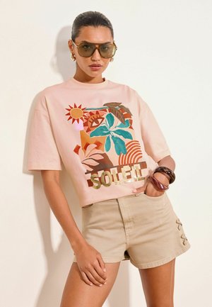 Frau trägt große Sonnenbrille, pinkes T-Shirt mit Sonnen- und Blätter-Motiv, beige Shorts und auffällige Armbänder, posiert vor einer schlichten Wand.