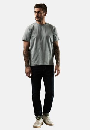 Lichtgrijze t-shirt met korte mouwen, ronde hals, gecombineerd met donkere jeans en sneakers. Bevat tatoeages op de armen en een neutrale achtergrond.