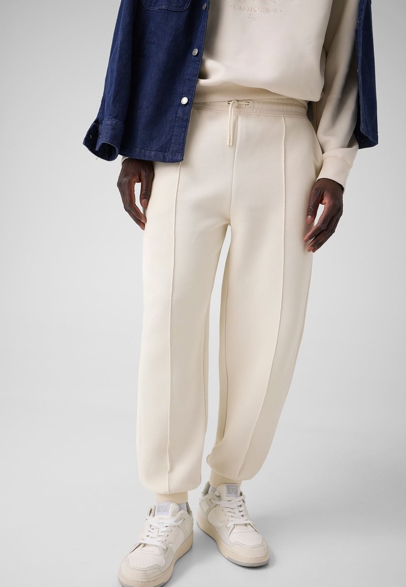 Personne portant un pantalon de jogging crème, un sweat crème, une surchemise bleue et des baskets blanches se tient devant un fond clair uni.