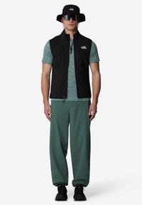 Black zip-up vest over a teal shirt, olive green pants, and black shoes. Accessories include a black bucket hat and large sunglasses.