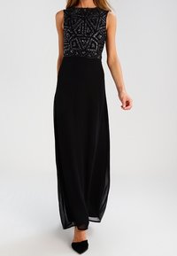 Kvinna i en ärmlös svart aftonklänning med geometriskt pärlade bodice och flödande chiffongkjol, matchad med svarta spetsskor.