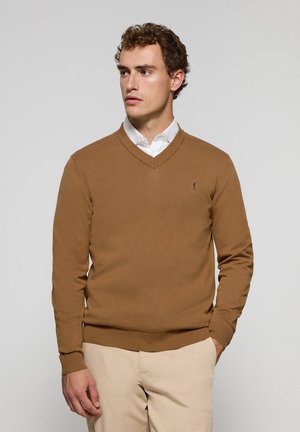 Brown v-neck sweater in knit fabric with ribbed cuffs and hem. Features a small logo on the chest. Worn over a white collared shirt.