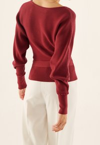 Burgunderfarbenes geripptes Sweatshirt mit weitem Bootsausschnitt und Puffärmeln, ausgestattet mit eng anliegenden Bündchen und einem Taillenakzentband. Trägt man zu cremefarbenen Hosen.