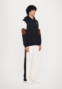 Hooded zip-up sweatshirt in navy with white and brown accents. Pairs with cream wide-leg pants. Features front pockets and logo detail.