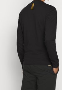 Sudadera negra de manga larga con acabado texturizado, que presenta un detalle de raya amarilla en la parte trasera del cuello y un corte relajado.