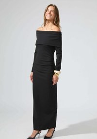 Robe longue noire décolletée, à manches longues, silhouette élégante, texture du tissu lisse, présentée avec un bracelet en or et des talons noirs.