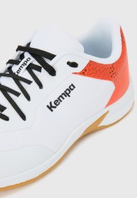 White athletic shoe with black laces, "Kempa" logo, textured orange accent, and gum sole, featuring a geometric pattern on the heel.