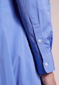 Ljusblå långärmad skjorta med manschett som har två vita knappar och sömnadsdetaljer på ärmen, bärs på en persons arm.