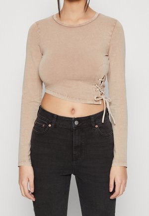 Frau trägt ein beiges, kurzes Langarmoberteil mit seitlicher Schnürung und hoch taillierte schwarze Jeans, steht vor einem einfarbigen Hintergrund.