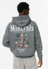 Persona che indossa un hoodie grigio con la scritta "Minerals" e illustrazioni colorate di minerali sulla schiena, abbinato a jeans chiari.