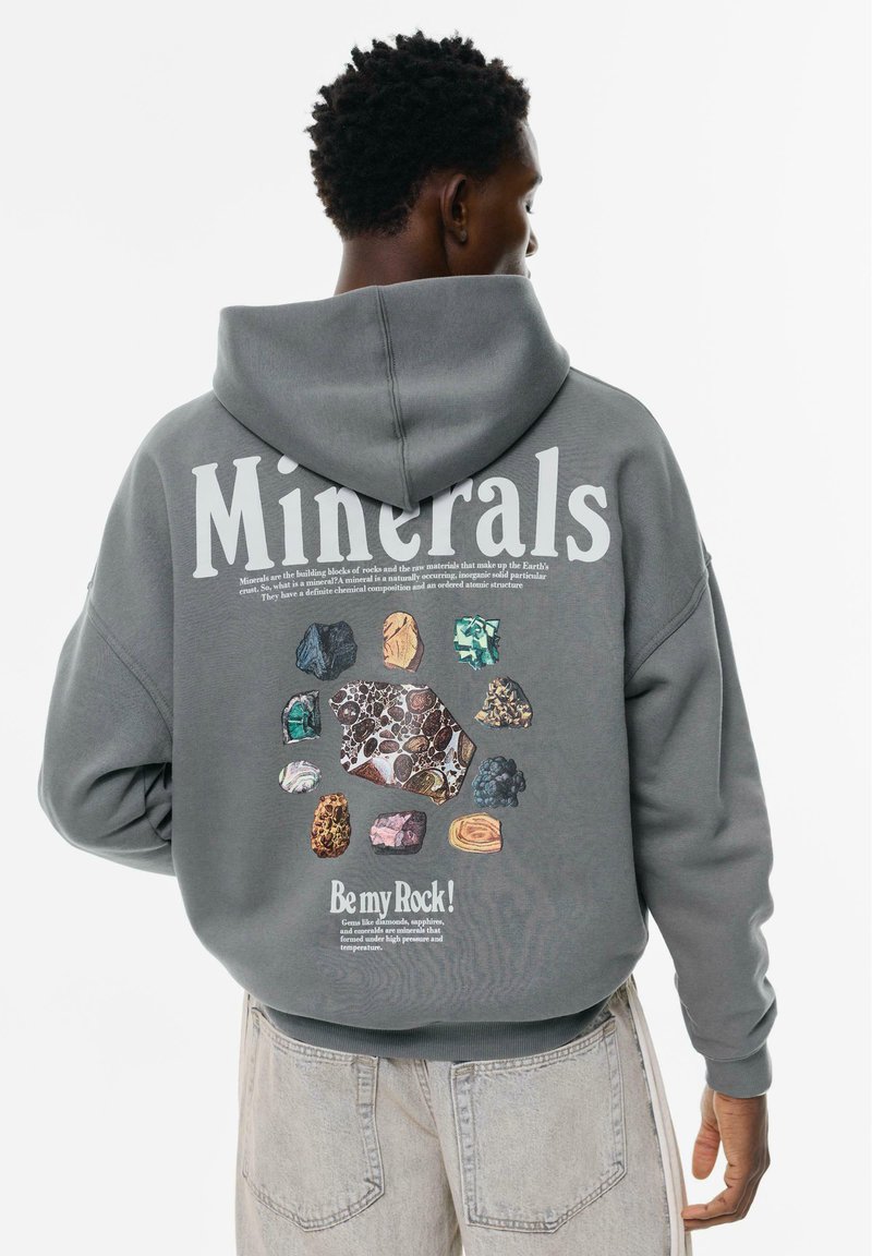 Persona che indossa un hoodie grigio con la scritta "Minerals" e illustrazioni colorate di minerali sulla schiena, abbinato a jeans chiari.