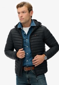 Sort pufferjakke med hætte, quillet design og rød accent. Båret over en denimskjorte, med et brunt bælte og armbåndsur.