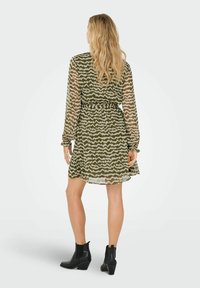 Vrouw met lang blond haar, gekleed in een gestreepte groen en beige lange mouwen jurk en zwarte enkellaarsjes, staand met haar rug naar de kijker.