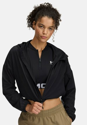 Zwarte cropped jack van lichtgewicht materiaal met een capuchon, voorzien van een rits aan de voorkant en elastische manchetten, gecombineerd met een zwarte top en tan-kleurige broek.