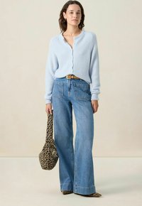 Femme portant un cardigan à boutons bleu clair, un jean bleu à jambes larges, une ceinture marron, tenant un sac à imprimé léopard et portant des chaussures à imprimé animalier à bouts pointus.