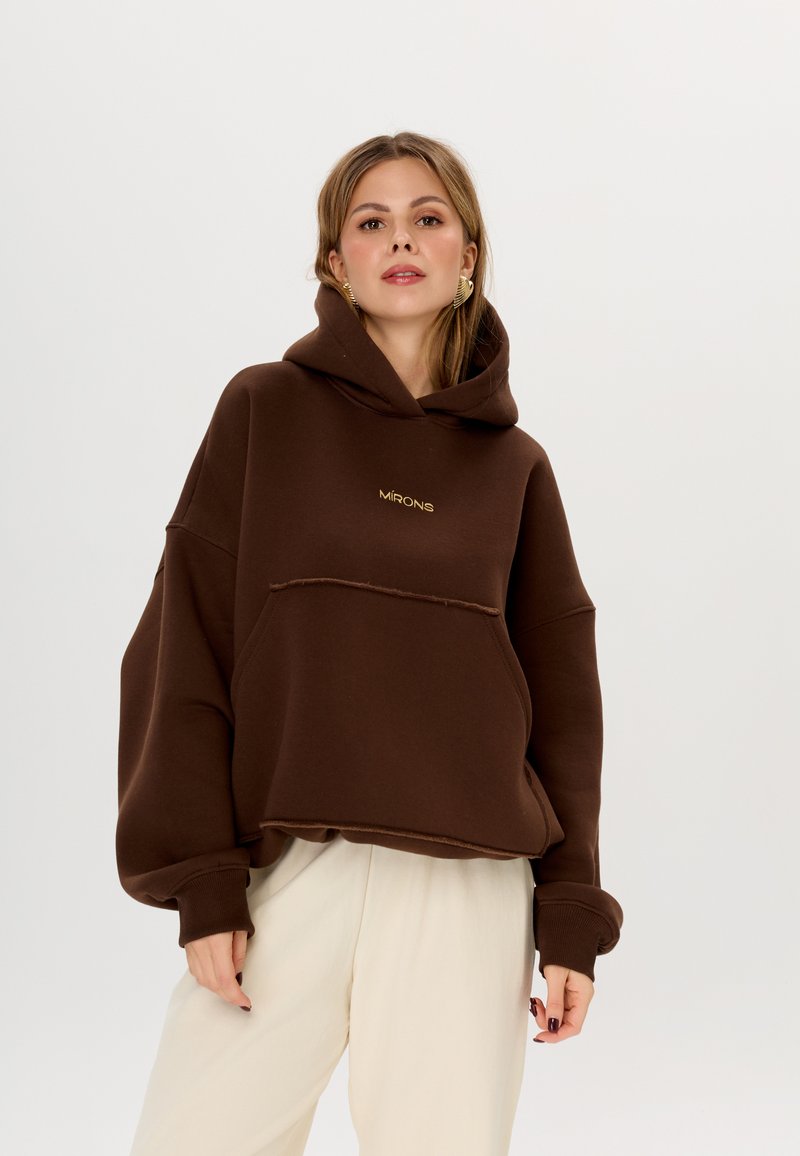 Felpa oversize marrone con tasca frontale e orlo grezzo, caratterizzata da un logo dorato "MIRONS" sul petto. Tessuto morbido e design con cappuccio.
