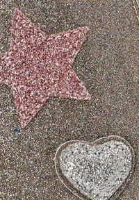 La superficie brillante presenta una estrella rosa y un corazón plateado, ambos con costuras delineadas. El material tiene una textura con elementos reflectantes y brillantes.
