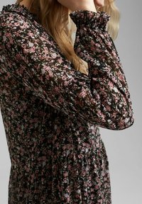 Vestido negro floreado con mangas largas y abullonadas, y puños con volantes. Presenta pequeñas flores rosas y blancas sobre una tela sheer y texturizada.