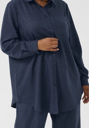 Detail osoby v nošené tmavě modré košili na knoflíky s náprsní kapsou a odpovídajících kalhotách na světlém pozadí.