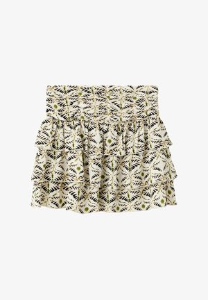 Kvinde iført beige hæklede haltertop, mønstret lagdelt mini nederdel og brune nitte-sandaler, stående mod en ensfarvet hvid baggrund.