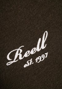 Dunkelbrauner Stoff mit weißer bestickter Schrift, die "Reell est. 1997" liest, mit einer glatten, leicht strukturierten Oberfläche.