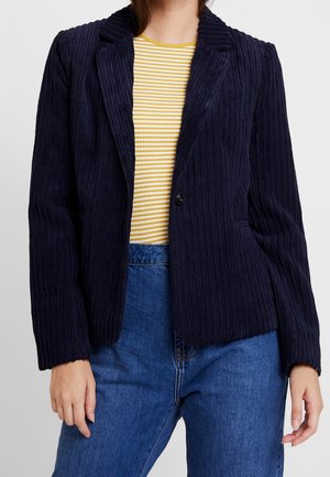 Woman wearing a dark navy ribbed blazer over a yellow and white striped top with blue jeans, standing with arms relaxed by sides.