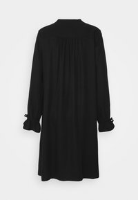 Robe noire à manches longues avec empiècement arrière froncé et poignets à volants noués par de petits nœuds, suspendue sur un fond blanc.