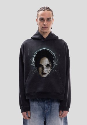 Person mit geflochtenem Haar trägt einen schwarzen Kapuzenpullover mit einer detaillierten Grafik eines Frauengesichts mit dunklen Augen und umgebendem elektrischen Design.
