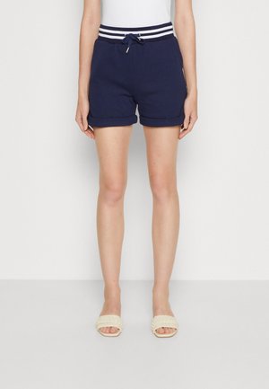 Kvinde iført marineblå shorts med striped talje og snor, kombineret med hvide åbne tåsandaler, stående mod en ensfarvet lys baggrund.