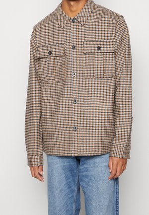 Mand iført brun og beige ternet skjorte med knapper og to brystlommer, kombineret med lyseblå denimjeans.