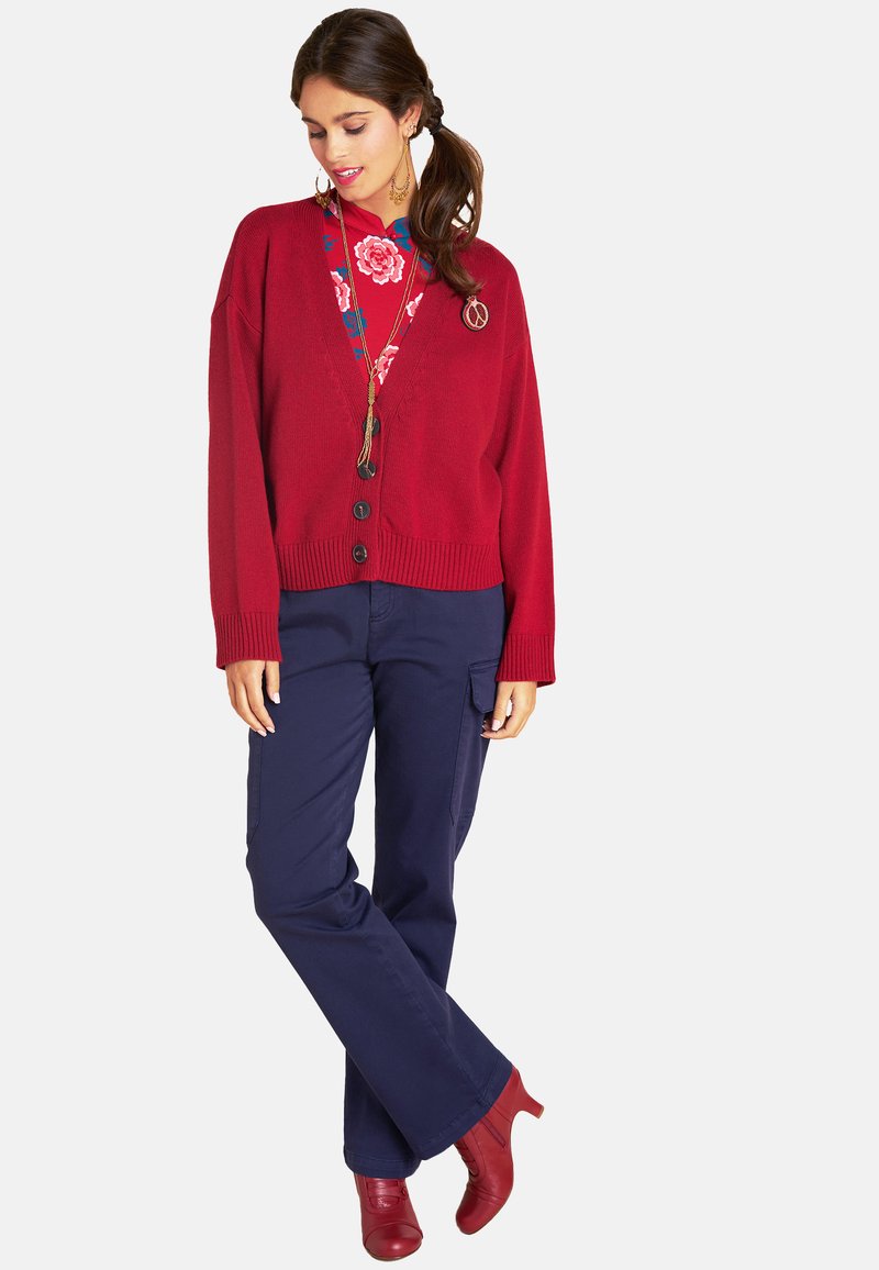 Roter gestrickter Cardigan mit schwarzen Knöpfen, kombiniert mit marineblauen Cargo-Hosen und roten High Heels. Blumendruck-Unterlage mit Akzenten.