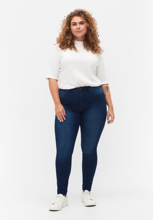 Skinny jeans in dark blue denim, featuring a high waist, five pockets, and subtle fading. Paired with a white ribbed t-shirt and light sneakers.
