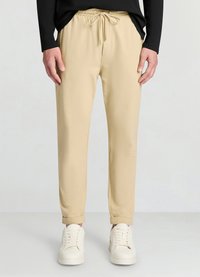 Beige Jogginghosen mit elastischem Bund und Kordelzug. Weicher Stoff mit glatter Textur, verjüngt sich zu den Bündchen hin. Kombiniert mit weißen Sneakers.